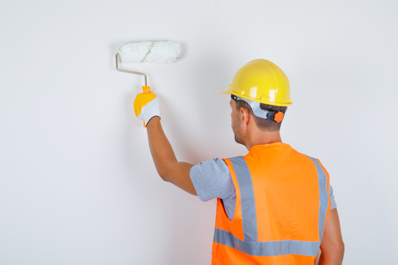 Male builder in uniform, helmet, gloves painting wall with roller , back view.